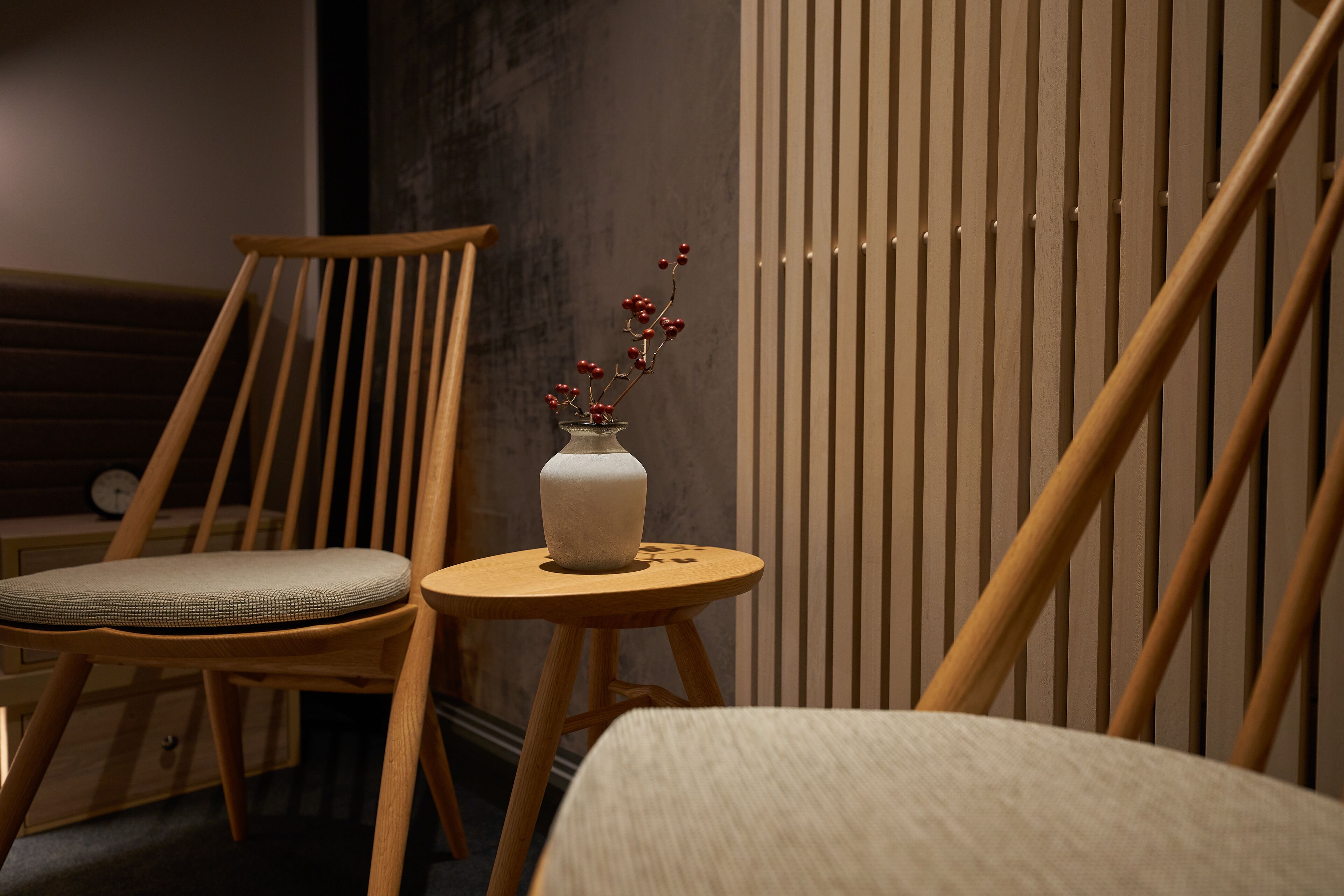 A wooden chair with a cushioned seat, a small round table with a white vase containing red berries, and a vertical wooden slat wall.