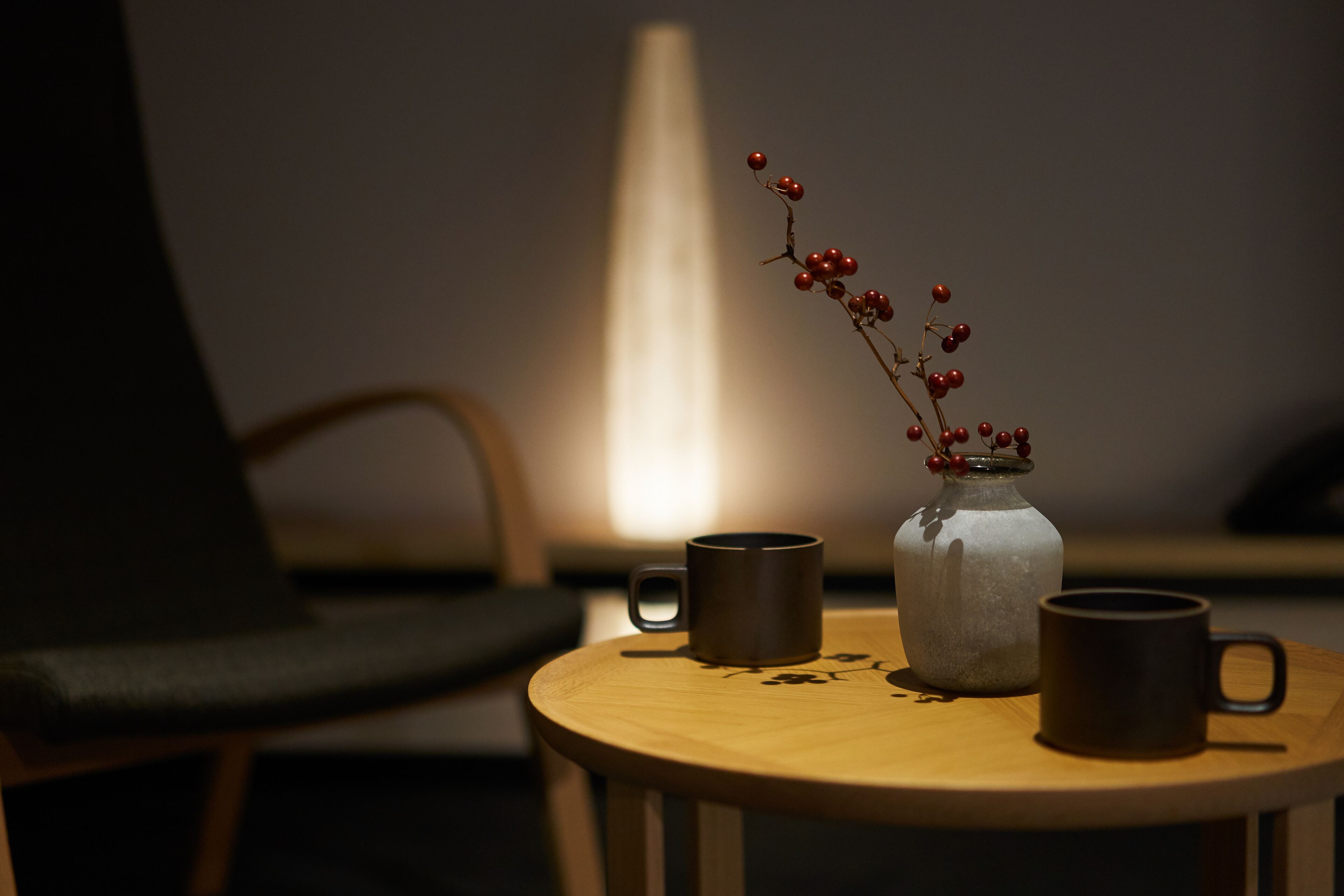 A round wooden table with two dark mugs and a clear glass jar with red berries.