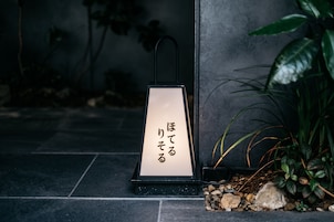 Un farol negro con caracteres japoneses sobre un suelo de piedra, rodeado de plantas.