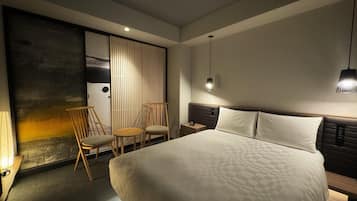 Una habitación de hotel moderna con una cama grande, dos sillas, una mesita de noche y un panel de persianas verticales en la pared.