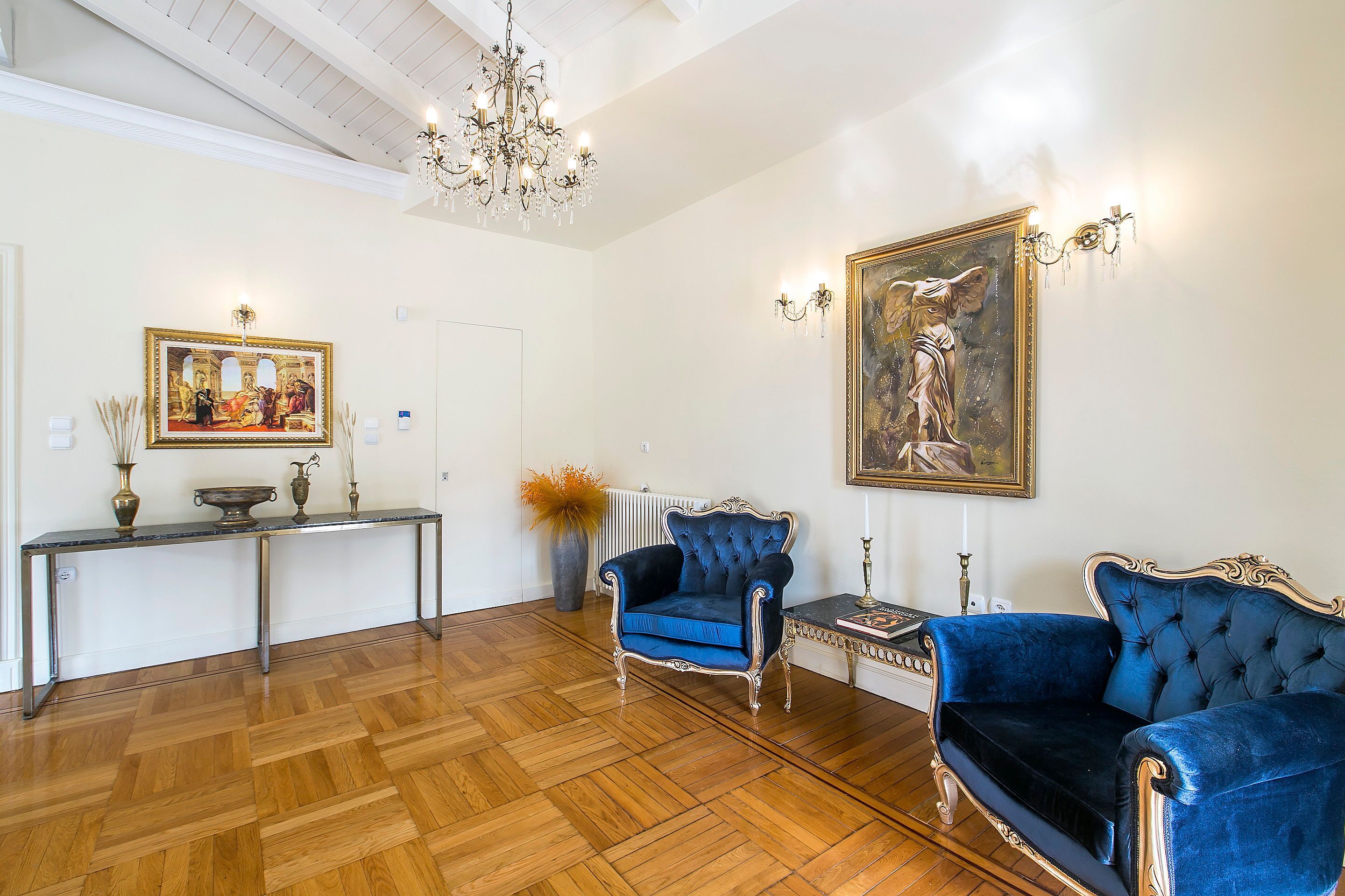 A spacious room with a wooden parquet floor, a blue velvet armchair, a matching sofa, a console table with decorative items, and a large framed painting on the wall.