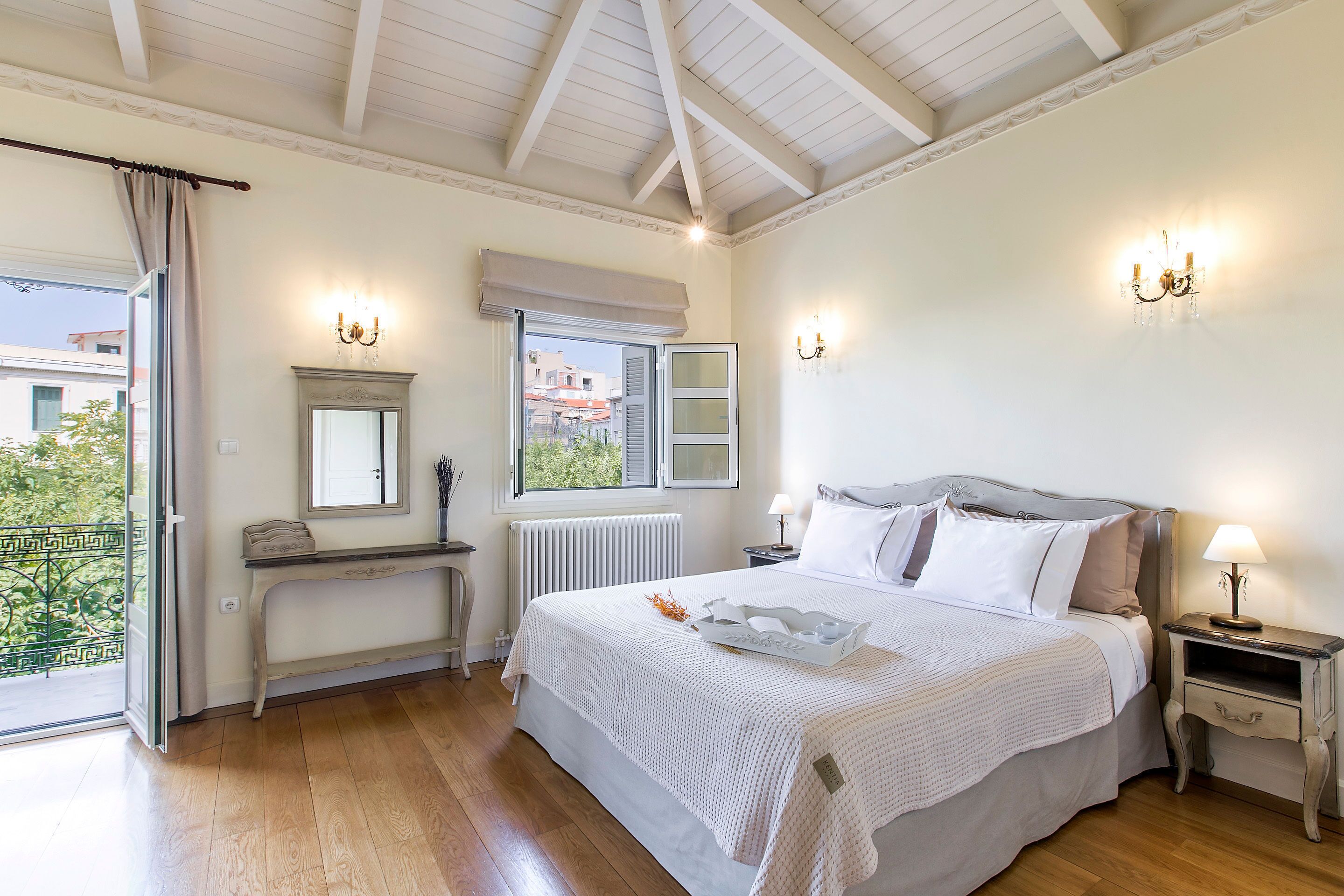 Ein Schlafzimmer mit einem großen Bett, Nachttischen, einem Schminktisch, einem Fenster mit Rollläden und einem Balkon mit Blick auf Gebäude.