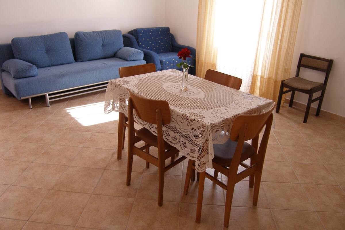 Ein Essbereich mit Tisch, bedeckt mit einer Spitzenleinentischdecke, umgeben von Holzstühlen. Eine blaue Couch lehnt an der Wand, daneben steht ein einzelner Sessel.