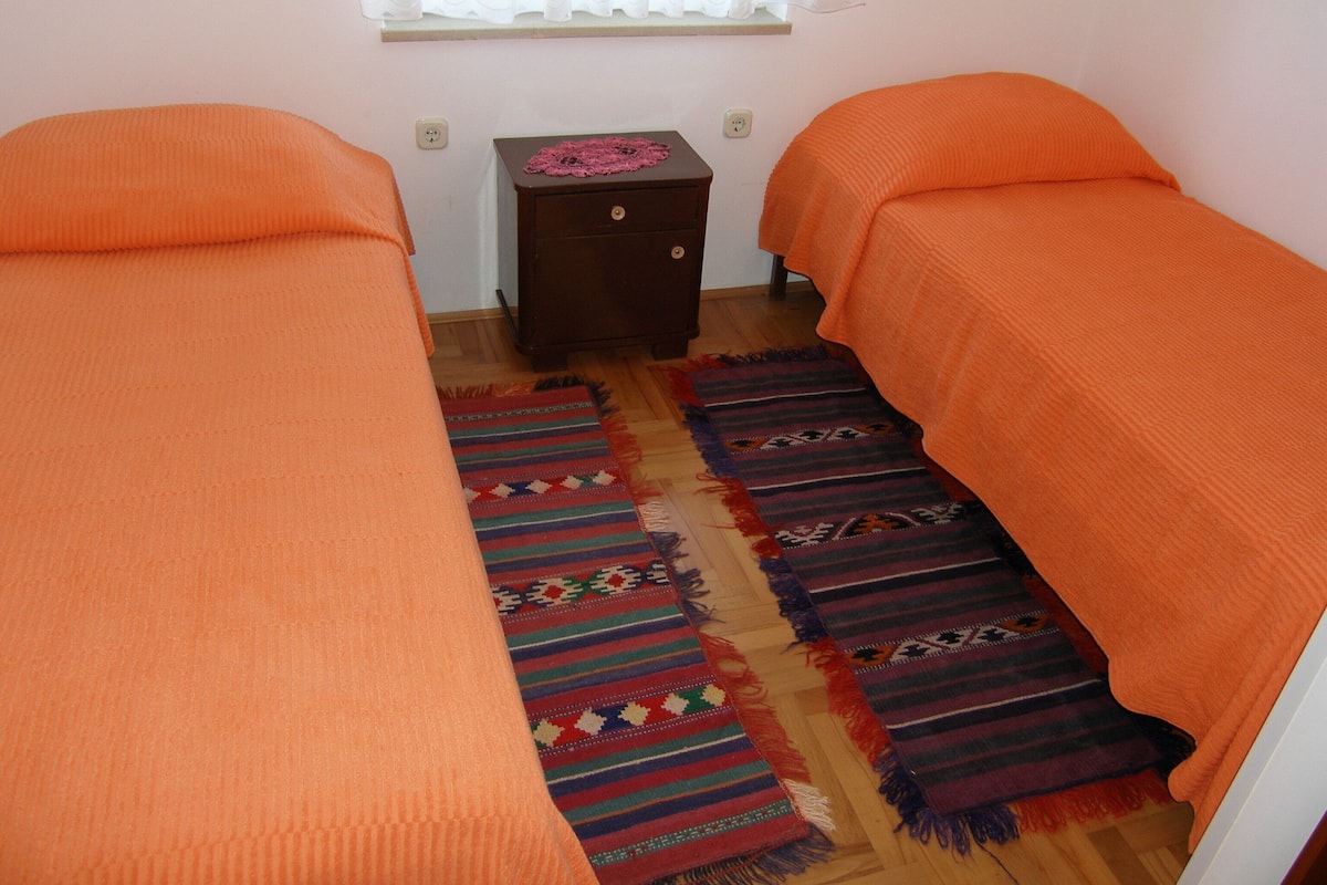 Ein Zimmer mit zwei Betten, einem kleinen Nachttisch aus Holz und einem Fenster mit Vorhängen.