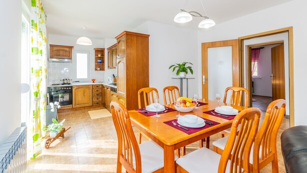 A dining area with a wooden table set for four, surrounded by wooden chairs. There is a kitchen in the background with wooden cabinets and a stove. - Family house with 2 apartments - Irma (Medulin)