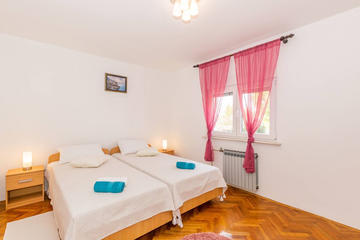 Ein Doppelbett mit weißer Bettwäsche und zwei blauen Handtüchern, ein Nachttisch mit Lampe sowie ein Fenster mit rosa Vorhängen.
