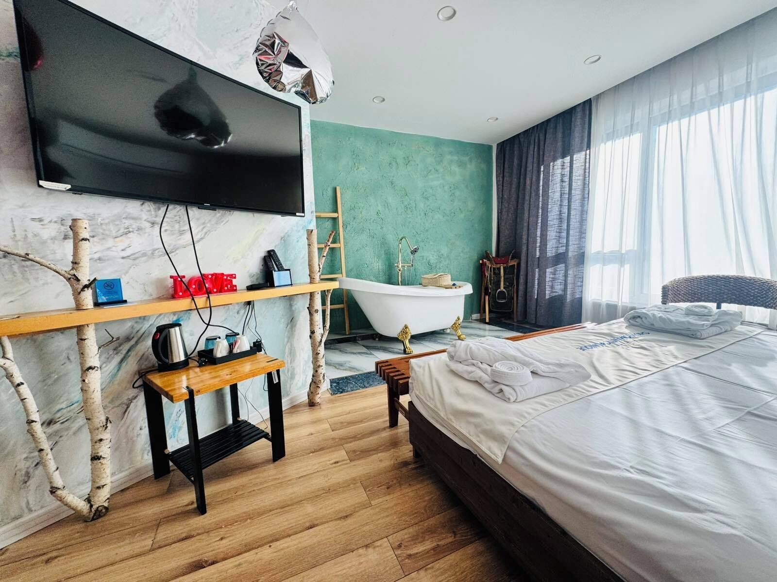 Une chambre à coucher moderne avec un grand lit, un téléviseur à écran plat fixé à un mur en marbre, une baignoire et une étagère en bois sur laquelle sont disposés divers objets.