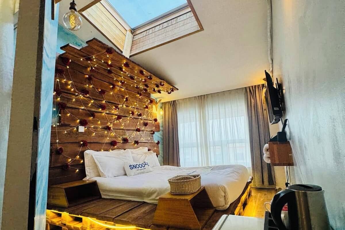A cozy room with a sloped ceiling, a wooden wall with string lights, two beds, a coffee maker, and a skylight.