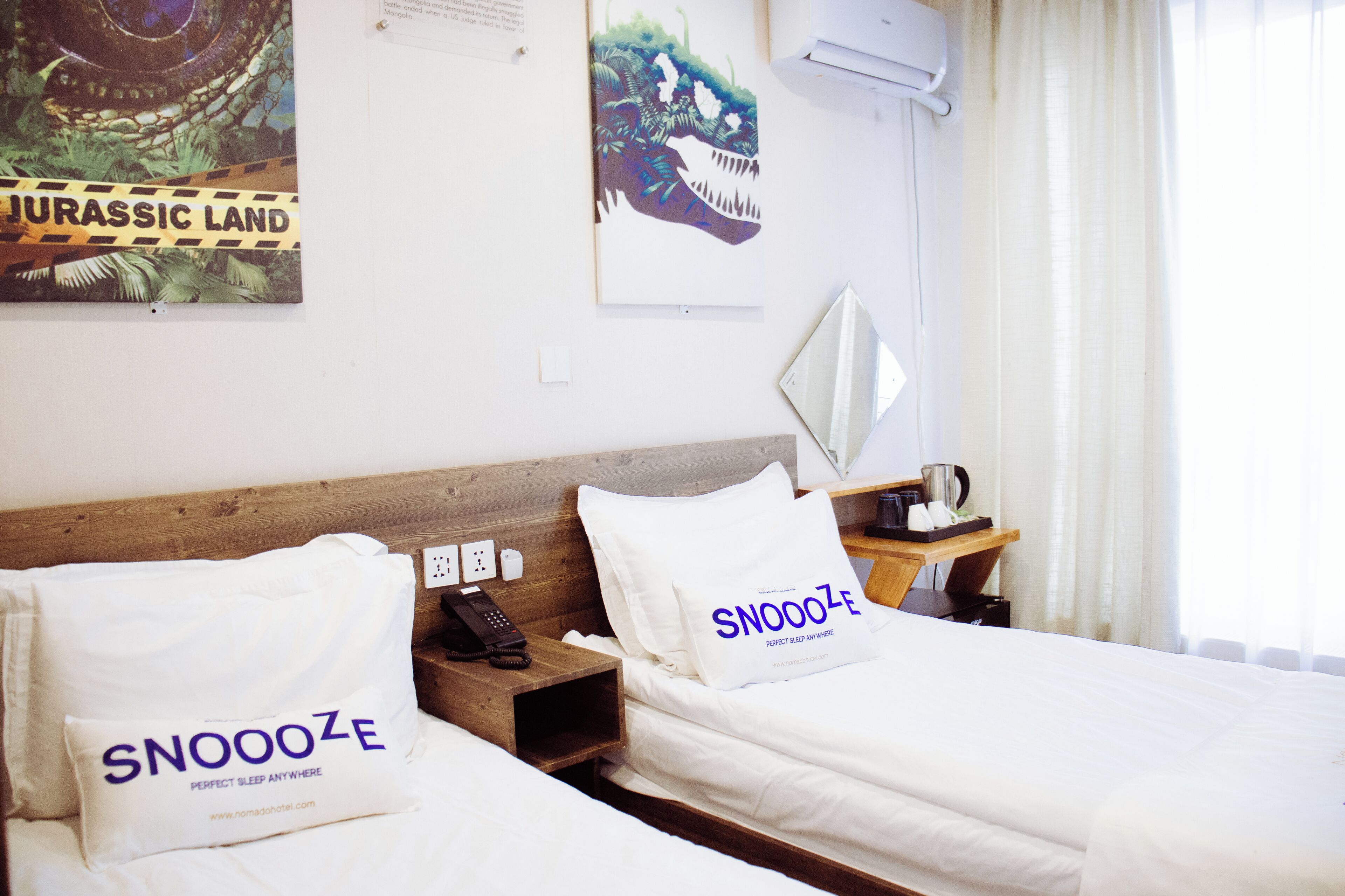 A hotel room with two beds, each with a pillow labeled 'SNOOZE' and a poster of a dinosaur on the wall.