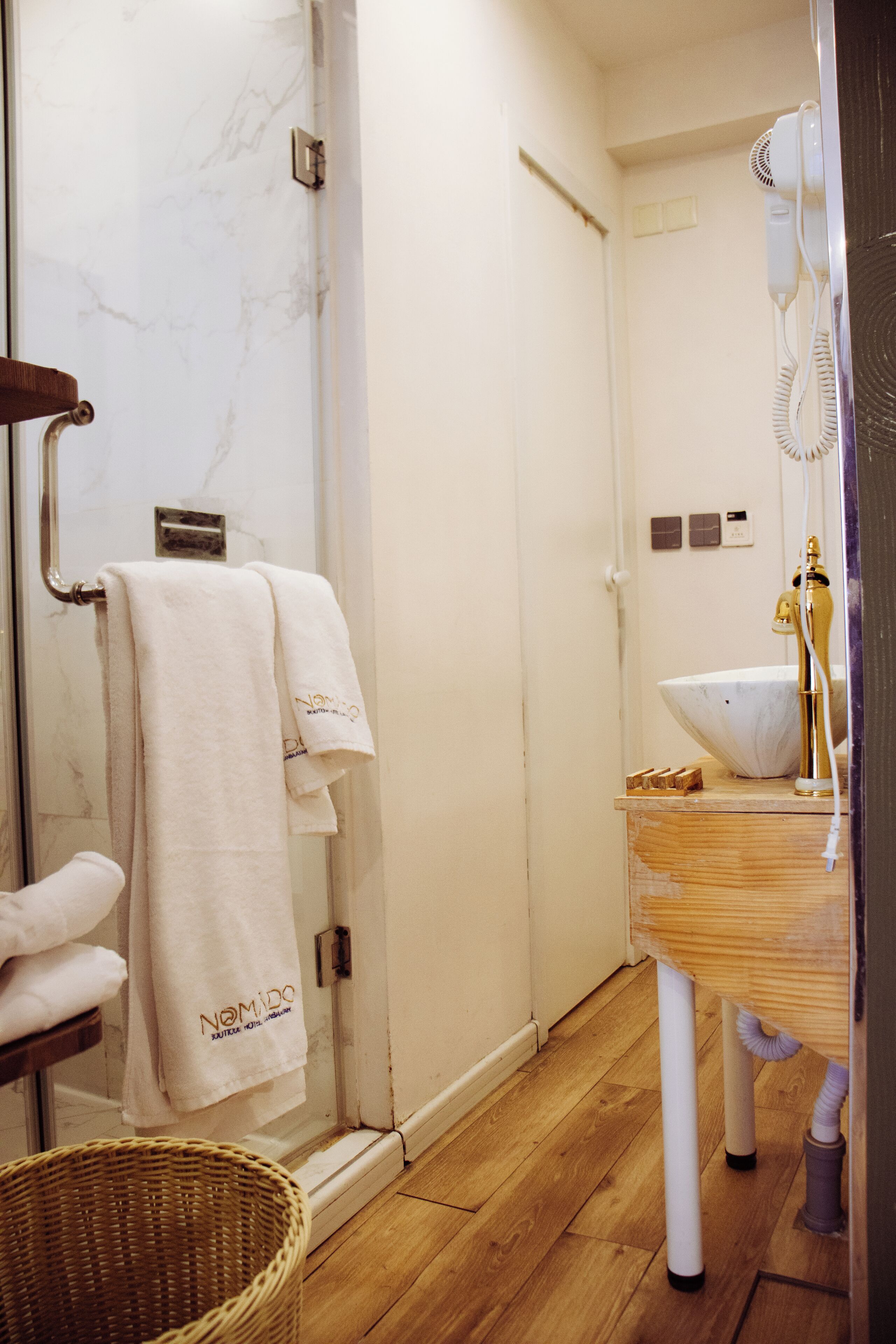 A bathroom with a glass shower enclosure, a wooden floor, a white sink with a gold faucet, and a towel rack with multiple towels.