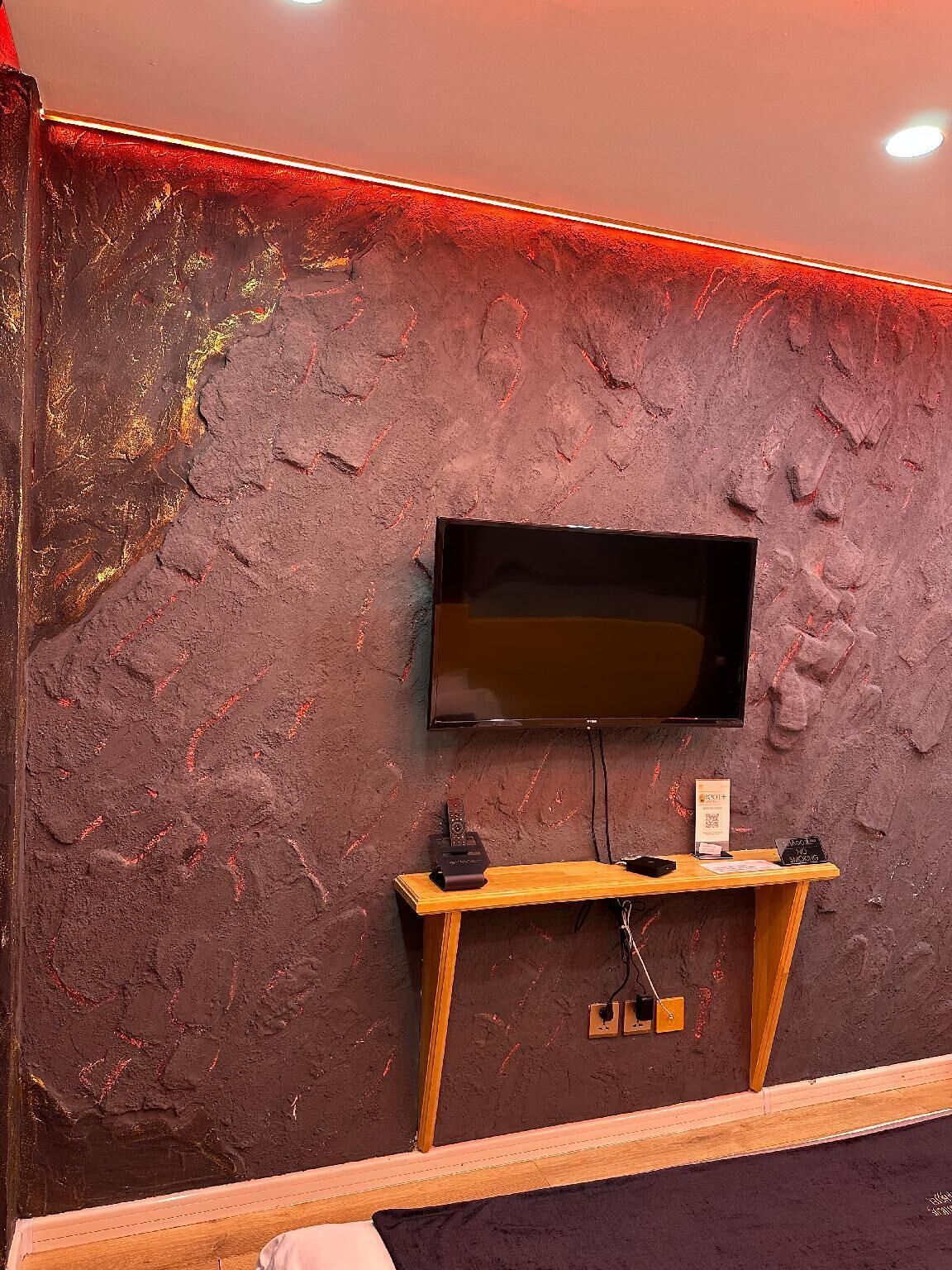 Un televisor montado en la pared con un estante de madera debajo, un enchufe y un cartel en la pared.