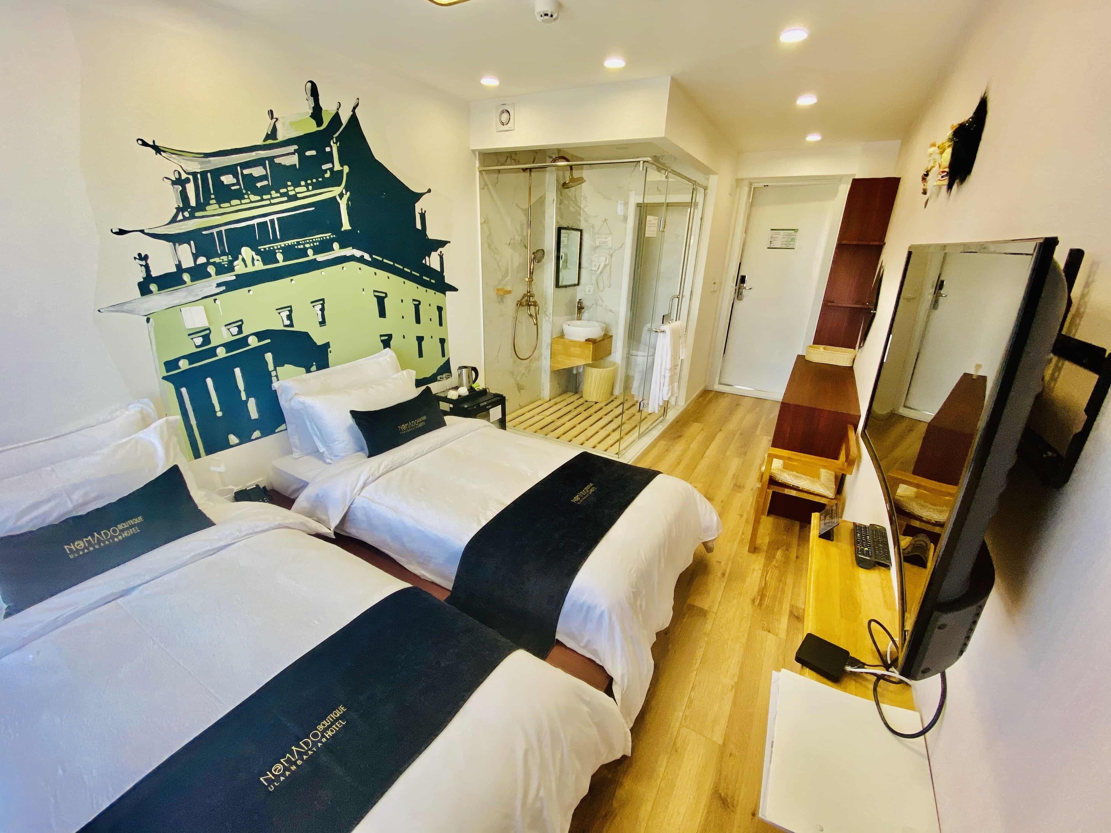 A hotel room with two beds, a bathroom with a glass shower, a wooden floor, and a TV on a stand.
