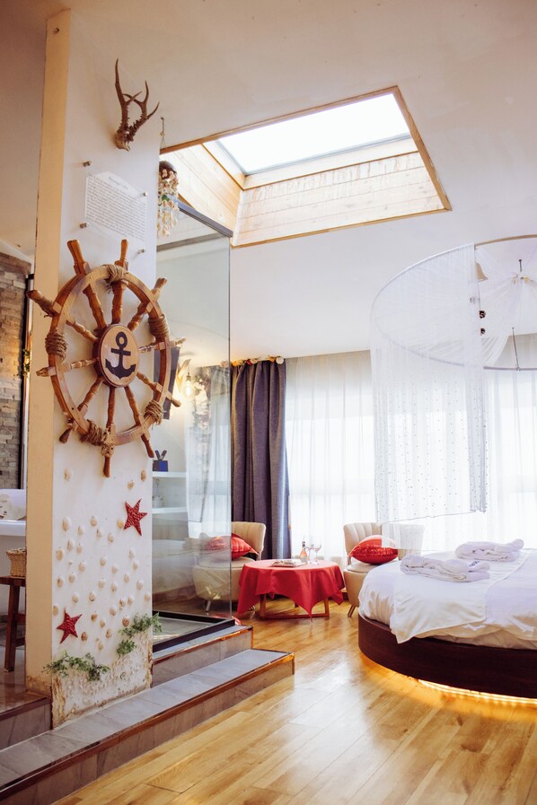 A hotel room with a bed, a large window, a ship's wheel decoration, and a staircase.