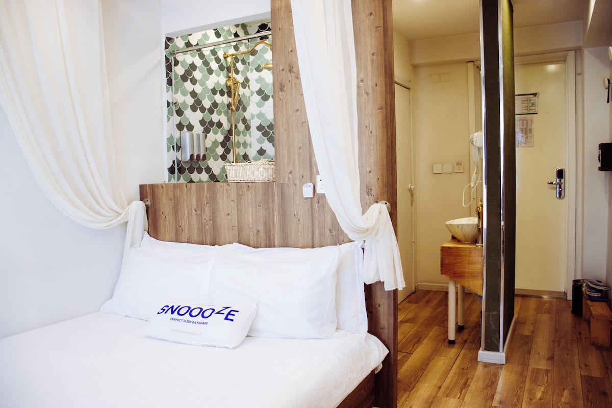 A hotel room with a bed, a wooden headboard, a white pillow with text, a shower area with a patterned glass door, and a wooden floor.