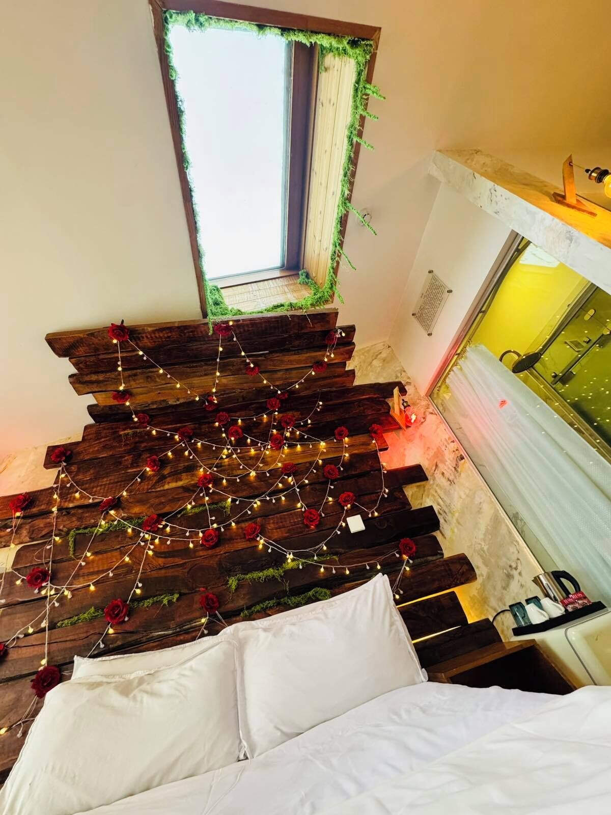 Una habitación con techo inclinado, una ventana con vistas, una cama con ropa blanca y una pared de tablones de madera con luces colgantes.