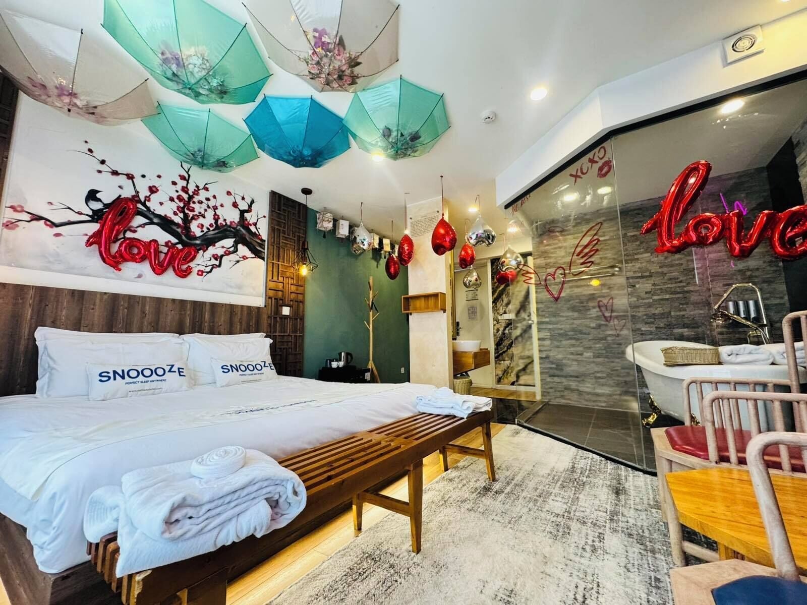 A hotel room with a bed, a bench, a mirror, and a bathroom visible through a glass partition.