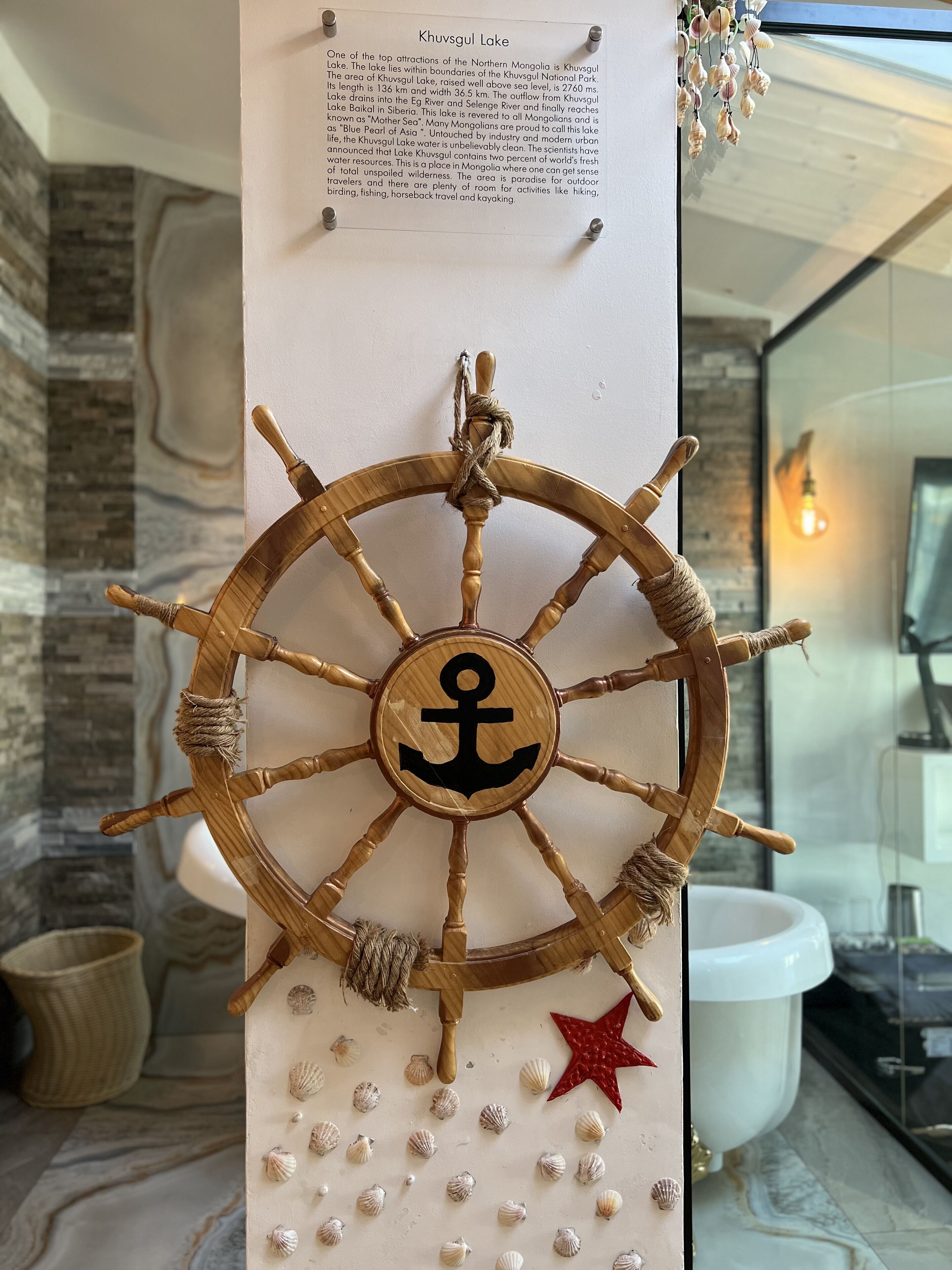 A wooden ship's wheel with an anchor symbol, surrounded by seashells and a star decoration, mounted on a wall with a text panel above.