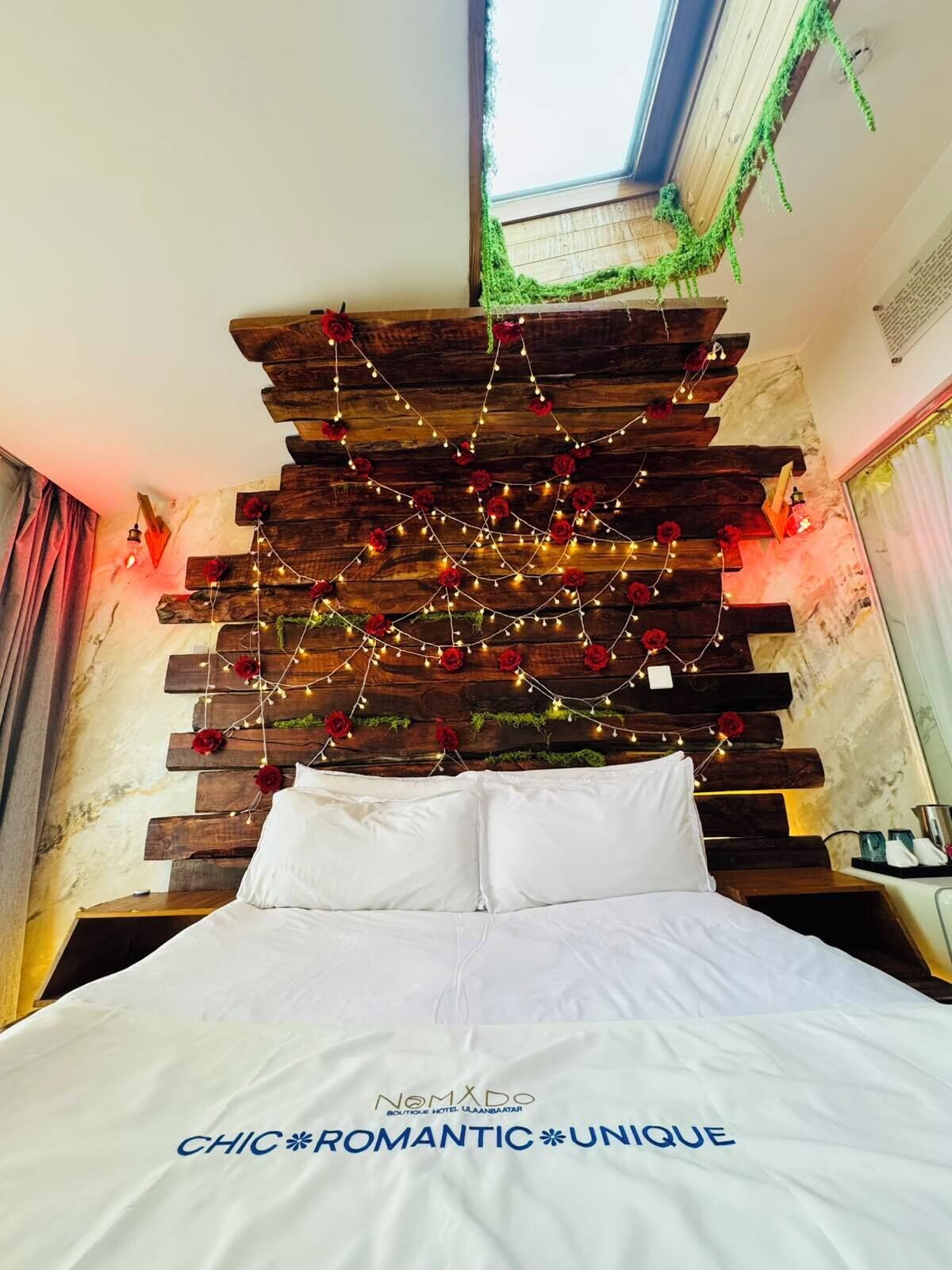 A bed with white bedding, a wooden headboard with string lights and red flowers, and a skylight above.