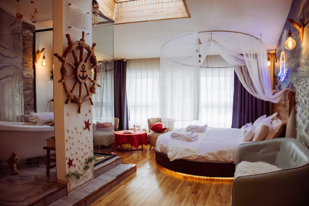 A hotel room with a bed, a sitting area with a table and chairs, a large window with curtains, and a ship's wheel decoration.