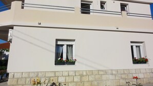 A two-story building with a flat roof, white exterior walls, and a stone base. There are two windows with curtains and flower boxes on the ground floor. - Apartments Cubela Beautiful Garden (Trogir)
