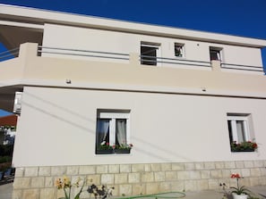 A two-story building with a flat roof, white exterior walls, and a stone base. There are two windows with curtains and flower boxes on the ground floor. - Apartments Cubela Beautiful Garden (Trogir)