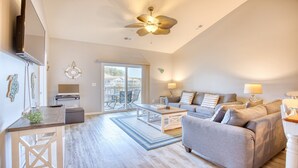 A spacious living room with a grey sofa, a coffee table, and a ceiling fan. There is a sliding glass door leading to an outdoor area with a patio table and chairs.
