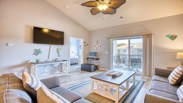 A spacious living room with a flat-screen TV, a ceiling fan, and a large sliding glass door leading to an outdoor patio.