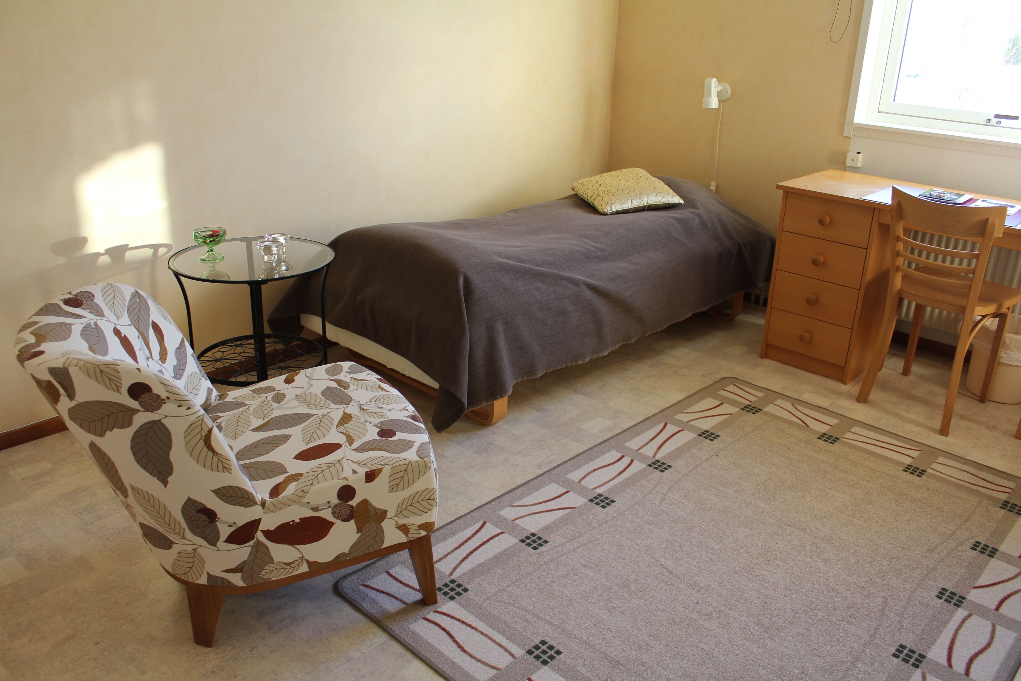 A small, neatly arranged bedroom with a bed, a desk, a chair, and a patterned armchair.