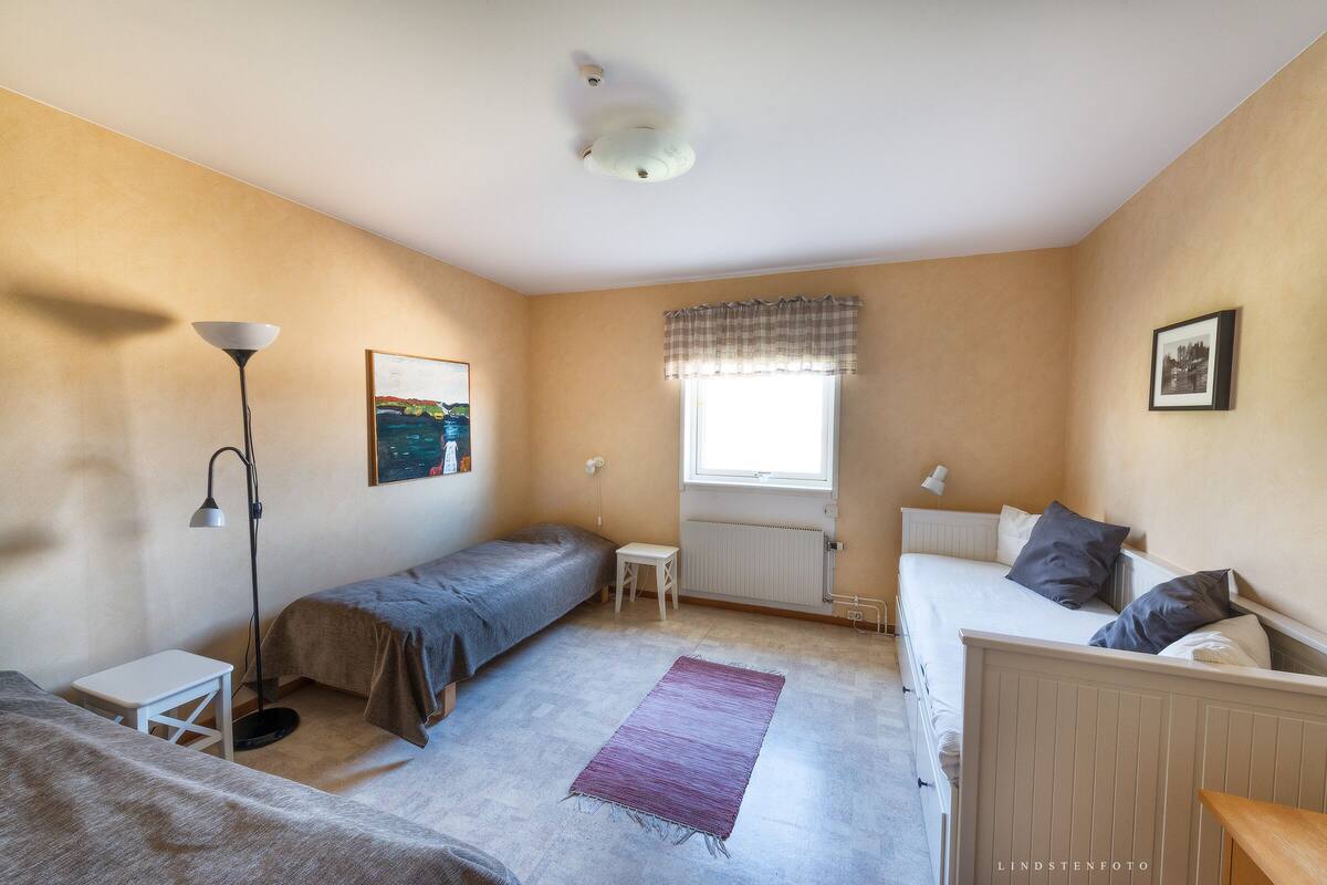 A room with two beds, a radiator, a window with curtains, a standing lamp, and a framed picture on the wall.