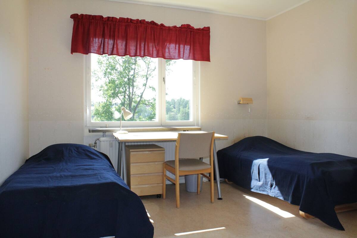 A dormitory room with two single beds, a desk, a chair, and a window with a red valance.