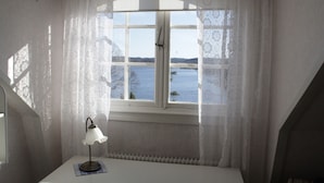 A room with a window overlooking a body of water, a lamp on a table, and a radiator below the window. - Liljeholmen Herrgård (Rimforsa)