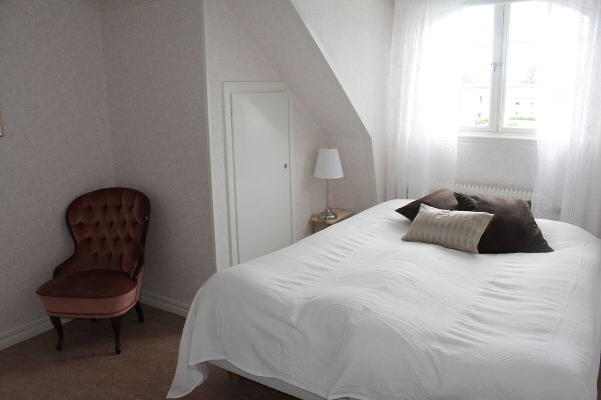 A bedroom with a large bed, a chair, a nightstand, and a window with curtains.
