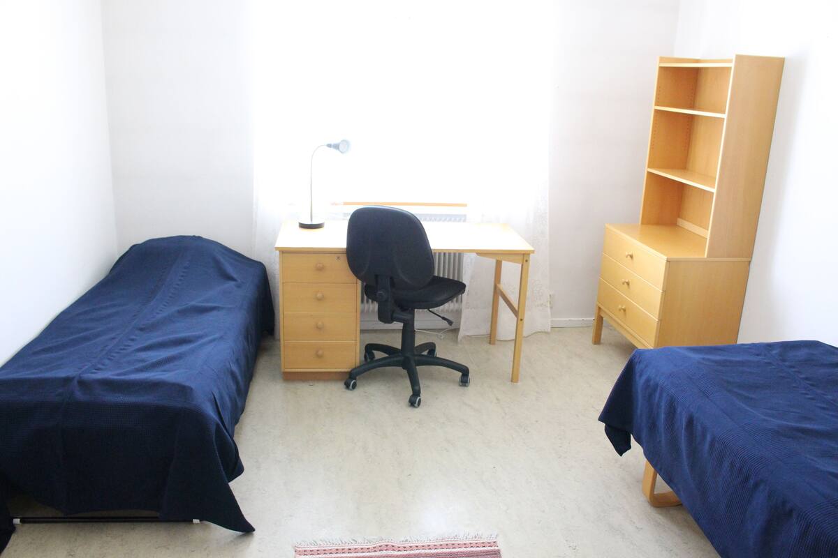 A room with two single beds, a desk with a chair, a radiator, and a bookshelf.