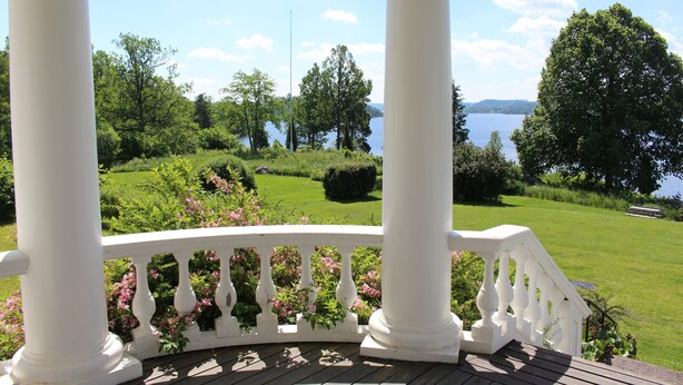 A wooden deck with white balustrades overlooking a lake and lush greenery. - Liljeholmen Herrgård (Rimforsa)