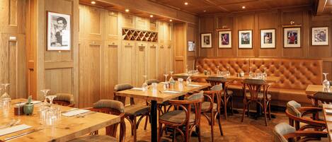 A restaurant with wooden paneling, multiple dining tables, cushioned chairs, and framed pictures on the walls.