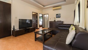 A living room with a dark brown leather sofa, a glass coffee table, and a flat-screen TV on a wooden stand. There is a door leading to another room and an air conditioning unit on the wall. - Orchid Paradise Homes OPV22 (Hua Hin)
