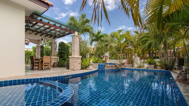 A swimming pool with blue tiles, surrounded by palm trees and a covered patio area.