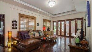 A spacious living room with a brown leather sofa, a glass coffee table, and a wooden cabinet. There are large windows with bamboo blinds, a chandelier, and a door leading outside.