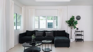 A modern living room with a black sectional sofa, two round coffee tables, a potted plant, a black clock on the wall, and a small black shelf unit. - Orchid Paradise Homes OPV02 (Hua Hin)