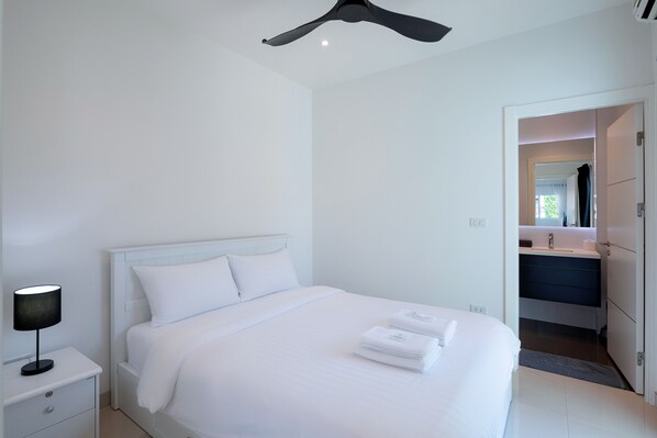 A clean, modern hotel room with a neatly made bed, a bedside table with a lamp, and a bathroom visible through an open door.