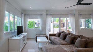 Un salon moderne avec un canapé, un téléviseur posé sur un meuble blanc et de grandes fenêtres ornées de rideaux.