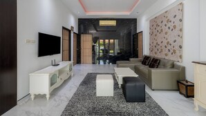 A modern living room with a flat-screen TV, a beige sofa, a white coffee table, and a black ottoman.