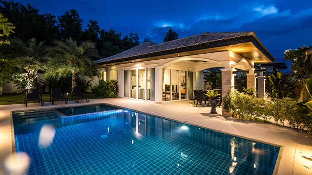 A swimming pool with a tiled deck, a covered patio area with seating, and lush greenery surrounding the property.