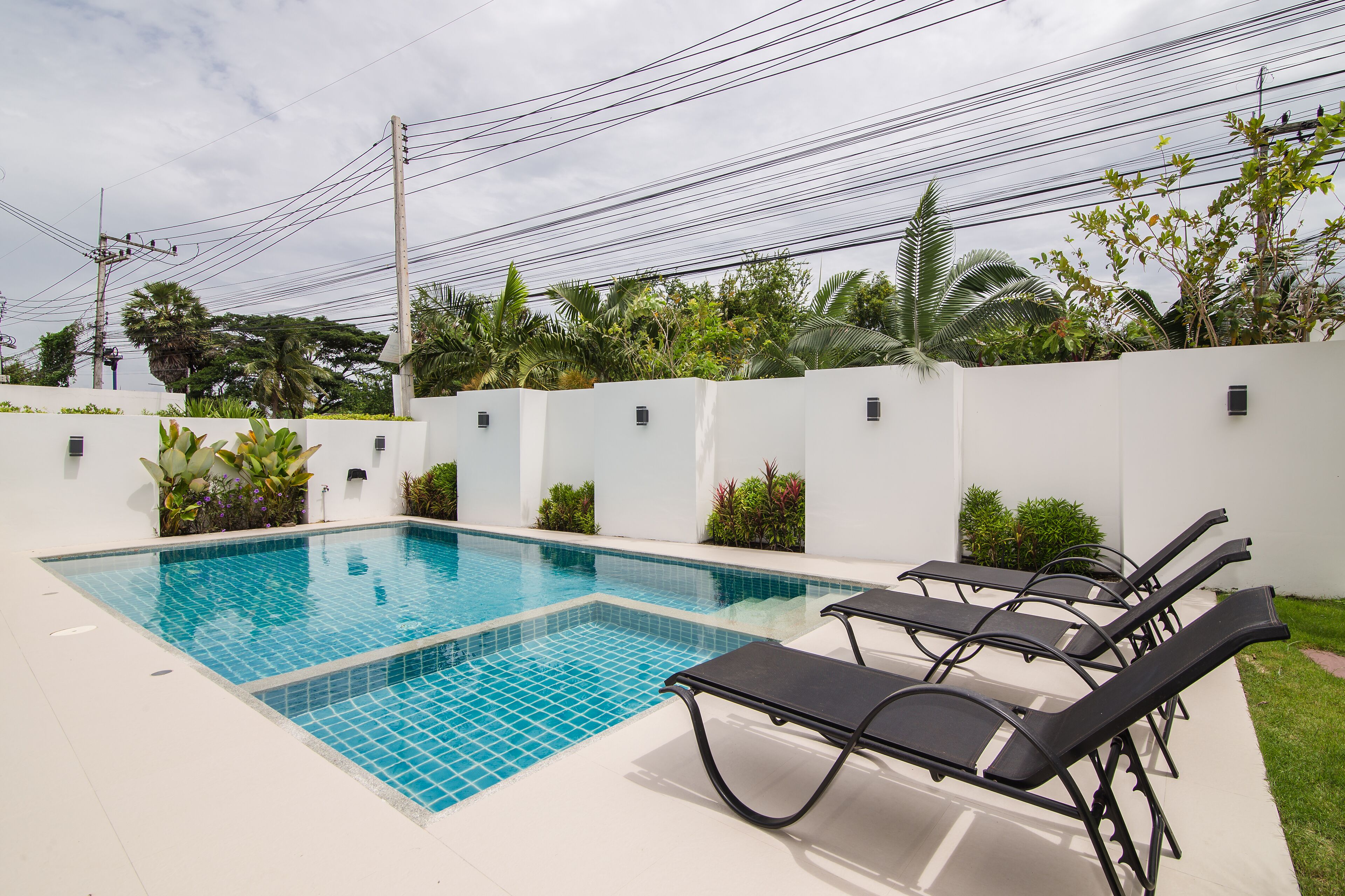 A swimming pool with tiled blue water, surrounded by white walls and green plants. There are black lounge chairs along the poolside.