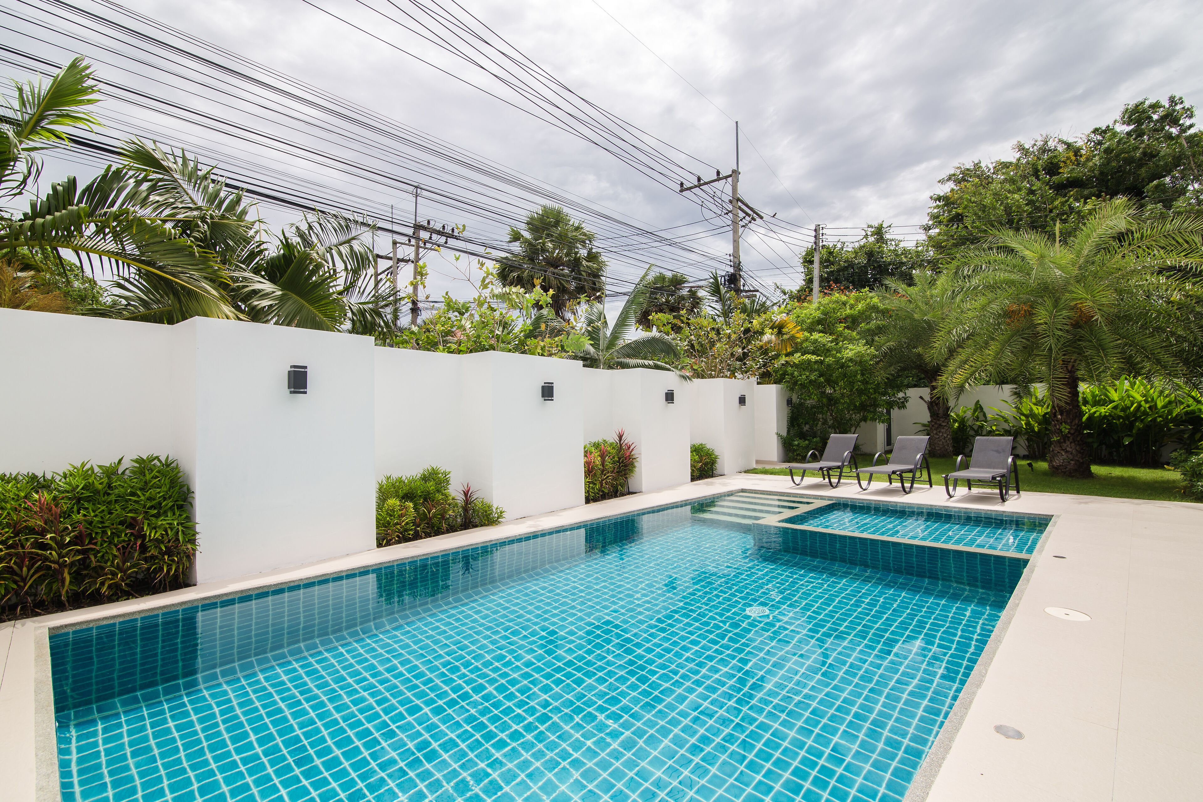 A rectangular swimming pool with a tiled blue surface, surrounded by a white wall and lush greenery.