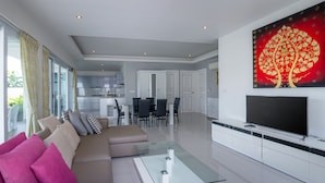 Ein modernes Wohnzimmer mit einem Glastisch, einem beigefarbenen Sofa mit pinken und grauen Kissen, einem Flachbildfernseher auf einer weißen Unterhaltungsanlage und einem roten Kunstwerk darüber.
