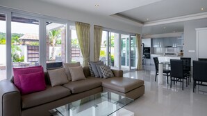 Ein modernes Wohnzimmer mit einem Glastisch, einem braunen Sofa mit Kissen und einem Essbereich mit einem weißen Tisch und schwarzen Stühlen.