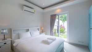 Ein Schlafzimmer mit einem großen Bett, weißer Bettwäsche, einem Nachttisch mit Lampe, einer Klimaanlage und Blick auf einen Garten durch eine Schiebetür.