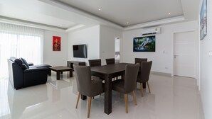 A modern living room with a dark wooden dining table, six chairs, a black sofa, and a flat-screen TV mounted on the wall. - Orchid Paradise Homes OPV401 (Hua Hin)