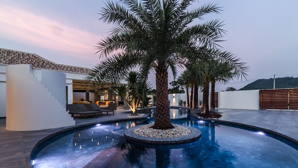 A modern outdoor pool area with a palm tree centerpiece, surrounded by a stone deck and a lounge area with seating. - Orchid Paradise Homes OPV301 (Hua Hin)